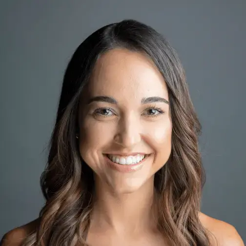 Une femme aux cheveux bruns souriant à l’appareil photo, sur un arrière-plan gris.