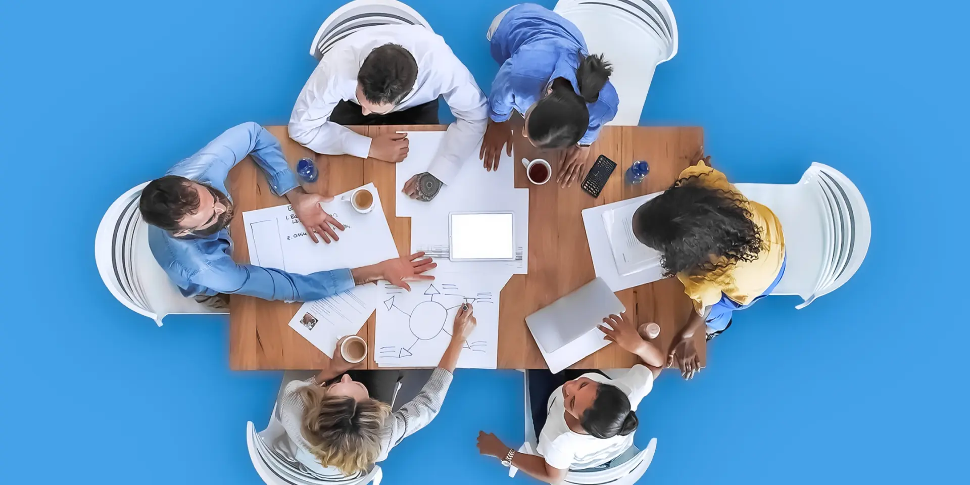 An image of an overhead view of 6 people around a wooden table, with papers scattered on the table. 