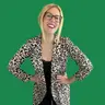 A smiling woman with blonde hair, glasses, and a leopard print cardigan poses with her hands on her hips in front of an olive green background.
