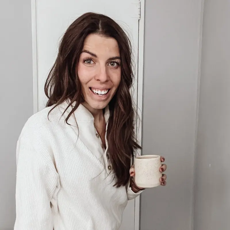 A woman with dark brown hair and a white sweater stands in front of a door smiling and holding a white coffee cup.