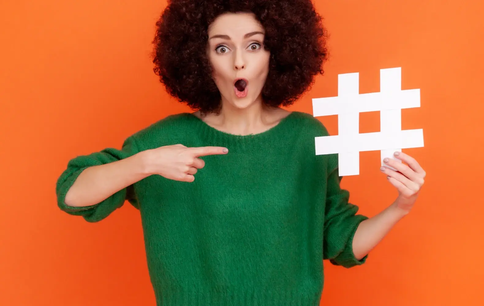 A person standing in front of an orange background pointing to a hashtag sign. 