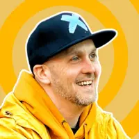 An image of Travis Brown, who is wearing a yellow sweatshirt with a black hat, set against a yellow background. 