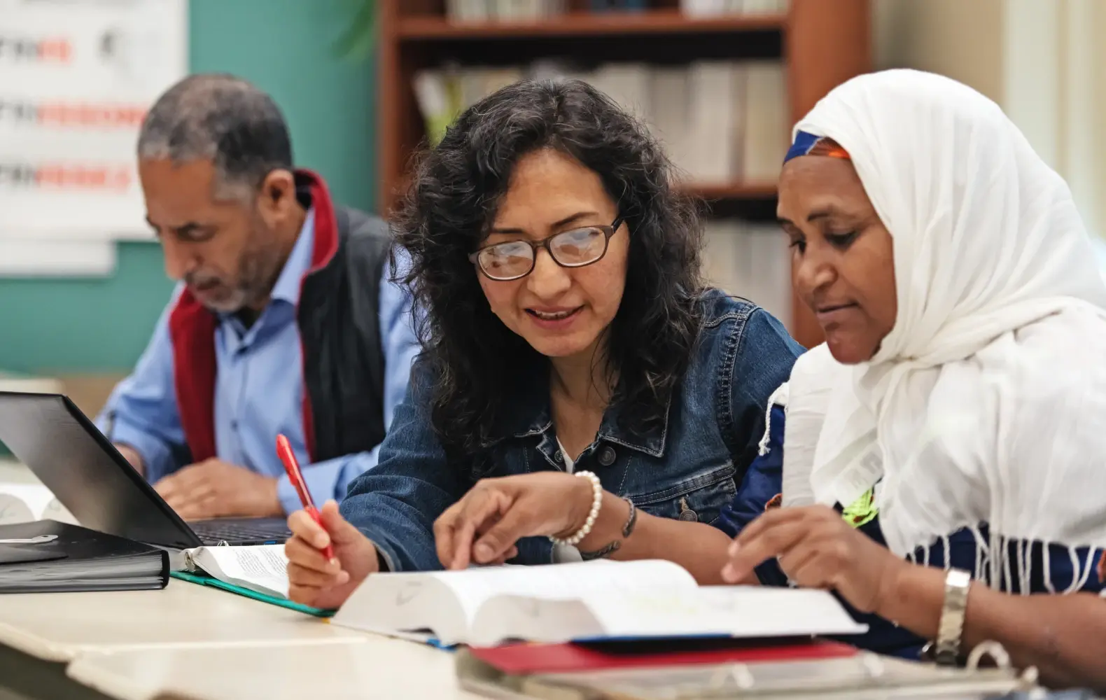 A female teacher with curly hair and glasses sits between two adult learners in an ESL classroom. The teacher reviews an open book with the learner in the foreground, a middle-aged woman wearing a hijab.