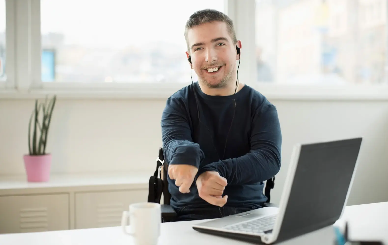 A young man in a wheelchair sits at a desk in front of an open laptop, wearing headphones and smiling. 