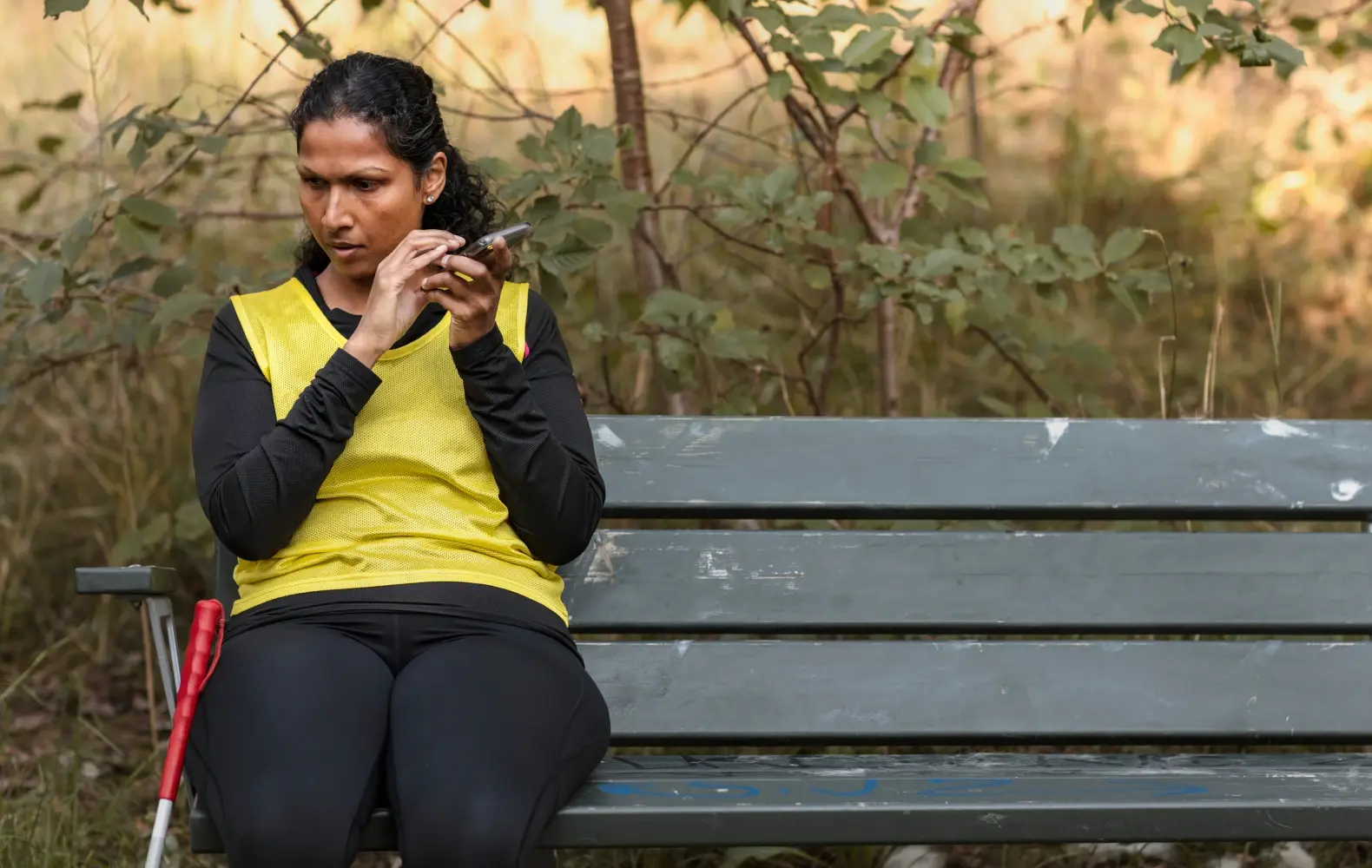 A blind woman with a cane sits on a park bench and holds her smartphone close to her ear, presumably to hear it narrate. 